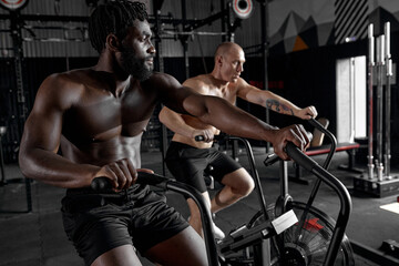 Fototapeta na wymiar Fit young diverse men using exercise bike at modern gym. Fitness male using bike for cardio workout at cross training club, side view on concentrated sporty male, focused on cardio workout