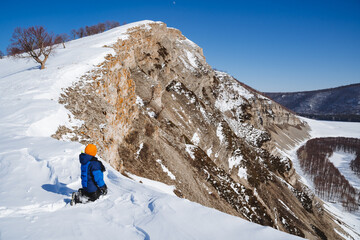 A little boy sits in the snow high in the mountains. A child travels through the winter snow climbing to the top of the mountain. Rocks against the blue sky.