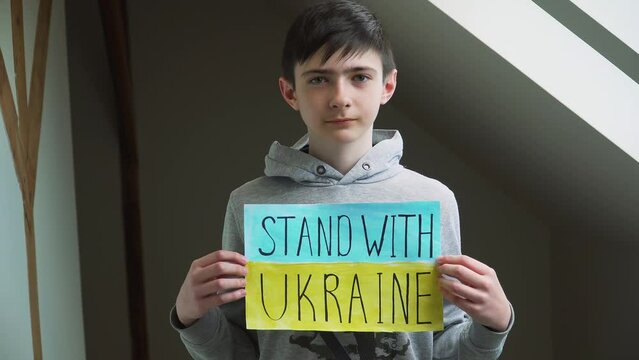 Portrait serious boy protesting armored conflict holding banner with inscription Stand with Ukraine at blue yellow flag. Crisis, peace, stop aggression in whole world