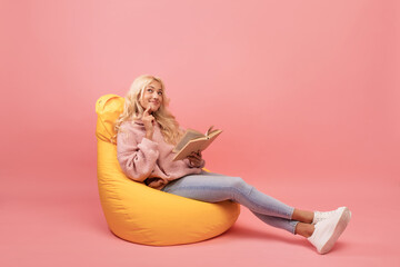 Dreamy lady reading book while sitting in comfortable beanbag chair over pink background, copy space