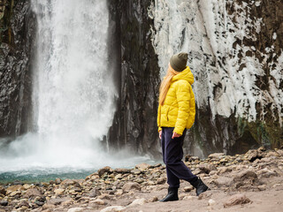 A woman in a yellow jacket looks at a stormy icy waterfall flowing down wet rocks on a cold November day.