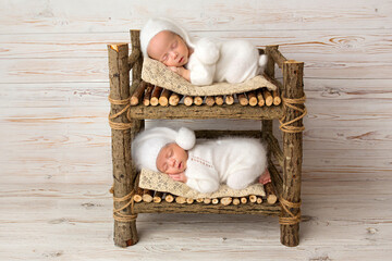 Tiny newborn twin boys in white bodysuits against a light wood background. Newborn twins sleep on a bunk wooden bed. The boys are dressed in white caps. Professional studio photography. - obrazy, fototapety, plakaty