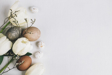 Stylish Easter eggs and tulips border on rustic white wooden background, flat lay with space for text. Happy Easter! Natural dyed eggs and spring flowers composition. Greeting card