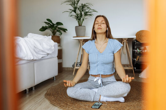 Trendy young woman wearing earphones meditating online and listening music on smartphone player app while sitting on floor at home,meditation app as part of mindfulness morning routine,peaceful mood.