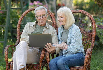 Modern senior spouses relaxing in wicker chairs in open air and using laptop computer, enjoying...
