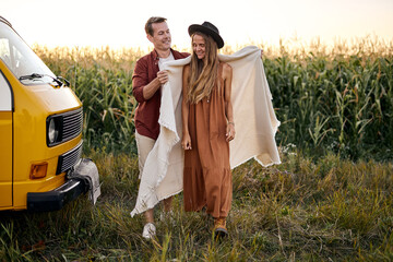Happy caucasian couple standing next to yellow minivan at sunset, travelling on summer vacation. Love and holiday concept. Cute man and woman in casual wear enjoy trip on van,female in plaid