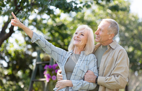 Happy Retired Couple Enjoying Time Together In Garden, Walking At Spring Day, Embracing And Woman Showing Something