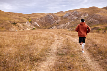 View from back on male running to mountains, dressed in sportive clothes, in red hoodie and black shorts, muscular body outdoors in nature. training,active lifestyle, health, sport concept