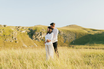 Man hugging his woman from behind while standing on the meadow with mountains on the background. Romantic moment, summer relaxing together concept.