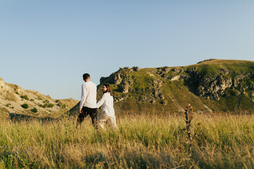 Couple holding hands and walking on the grass on a background of mountains and blue sky.