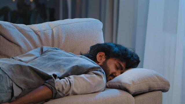 Exhausted syndrome bad feeling tired arabian man indian bearded guy worker male at home falling down face on couch sofa late time night evening dark overworked sick person lack of energy after party