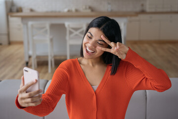 Young emotional influencer using mobile phone streaming video relaxing at home. Beautiful African American woman taking selfie, showing victory sign, communication online