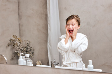 surprised teenage boy in bathrobe looking in mirror opened mouth in bathroom at home after cleaning...