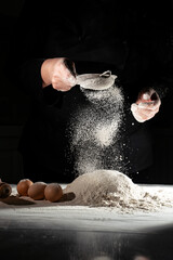 woman sifts flour through sieve onto table with eggs on black background. 