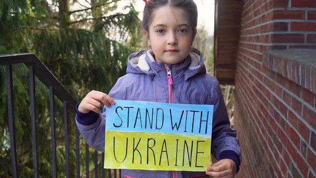Toddler girl kid homeless protesting armored conflict holding banner with inscription message text Stand with Ukraine on background of yellow blue flag. Crisis, peace, stop aggression in whole world