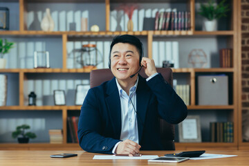 Asian businessman happy talking on video call in classic office, working at desk, using headset,...