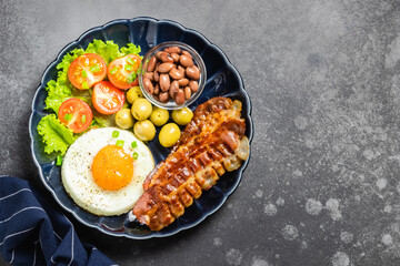 Traditional English breakfast, roast bacon fried egg, tomatoes, lettuce and olives on dark background. Top view, copy space.