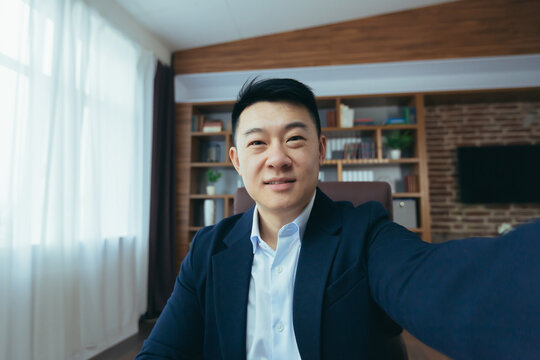 Cheerful and funny Asian businessman looks at the camera of a smartphone, talks on a video call, a man works in the office, waves a congratulatory gesture