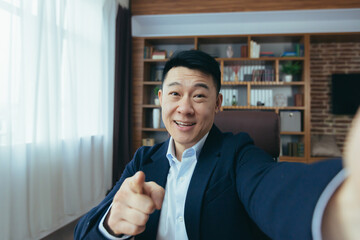 Cheerful and funny Asian businessman looks at the camera of a smartphone, talks on a video call, a...