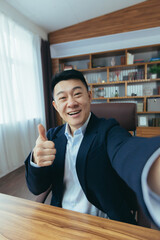 Cheerful and funny Asian businessman looks at the camera of a smartphone, talks on a video call, a man works in the office, waves a congratulatory gesture