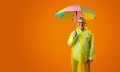 Senior man wearing yellow plastic overcoat and holding rainbow umbrella over yellow background