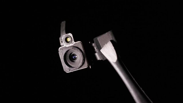 A close-up camera module appears from the darkness and disappears. Hidden camera on black background