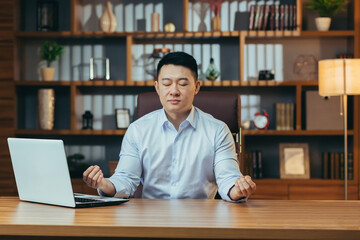 Successful businessman meditates sitting at a table in a classic office, Asian man in shirt relaxes, after working on laptop, man at work