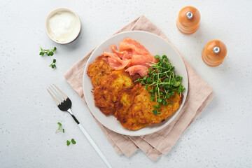 Potato pancakes. Fried homemade potato pancakes or latkes with cream, green onions, microgreens, red salmon and sauce in rustic plate on white table background. Rustic style. Healthy food. Top view.