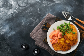 Potato pancakes. Fried homemade potato pancakes or latkes with cream, green onions, microgreens, red salmon and sauce in rustic plate on old black table background. Rustic style. Top view.
