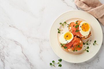 Sandwich with delicious toppings, smoked salmon, eggs, herbs and microgreens radish, black sesame seeds over white plate on white marble table background. Healthy open sandwich superfood. Top view.