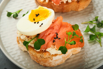 Sandwich with delicious toppings smoked salmon, eggs, herbs and microgreens radish, black sesame seeds over white plate on gray concrete table background. Healthy open sandwich superfood. Top view.