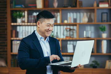 Happy asian freelance businessman working standing, holding laptop, smiling and rejoicing, at home...