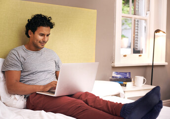 Doing some bedroom browsing. Shot of a young man sitting on his bed browsing on his laptop.
