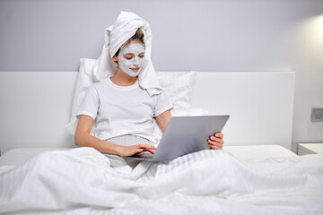 Young woman with clay facial mask using laptop while sitting on bed at home. Woman having face treatment at home lying on bed. Woman use beauty mask, at home alone. skin care, beauty concept