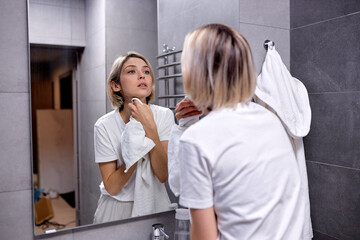 beauty, skin care and people concept. woman in hairband waping face with towel and looking to mirror at home in bathroom. short haired lady in casual domestic wear enjoy skin care, in the morning