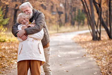 Excited Friendly Senior Elderly Couple In Coats Enjoy The Walk Together, Hugging, Laughing. Caucasian European Married Man And Woman In Love, During Autumn Season. Happiness, Lifestyle