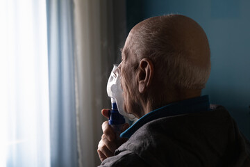 Back view angle headshot of an old man with asthma using a nebulizer in his room sitting on a bed