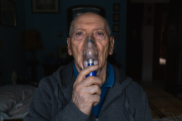Front view of an old man with asthma using a nebulizer in his room sitting on a bed