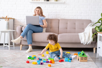 Naklejka premium Mother freelancer use laptop sits on couch, kid daughter plays with toys on floor. Working from home