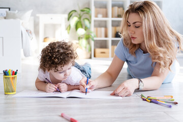 young mother and curly child girl drawing with colored markers lying on floor room at home