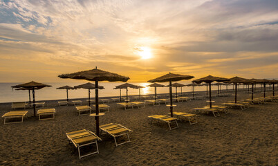  empty beach during beautiful sunrise or sunset with chaise loungues and nice umbrellas with blue sea, sun glow and amazing cloudy sky on thr background