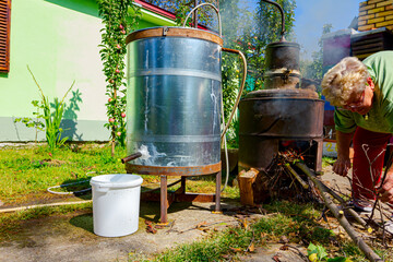 Obraz premium Woman is throwing firewood for maintaining fire under the distillation apparatus, it making domestic alcohol liquor