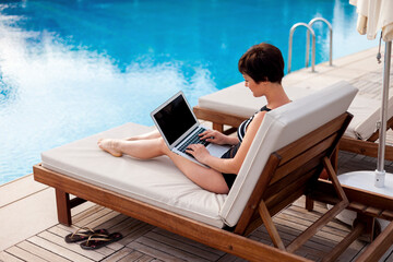 a woman in a swimsuit with a laptop lies on a sun lounger by the pool and looks at the monitor screen. work online. swimming pool on site
