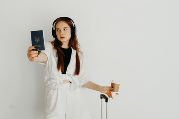 Naklejka premium Young woman with cup of coffee, headphones, and a suitcase showing her passport, on a white background. Travel concept. Woman is going to travel by plane
