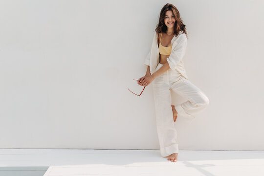 Full-length slender fair-skinned young woman smiles at camera while standing against white wall. Brunette with loose wavy hair in top, shirt and trousers. Happy weekend concept.