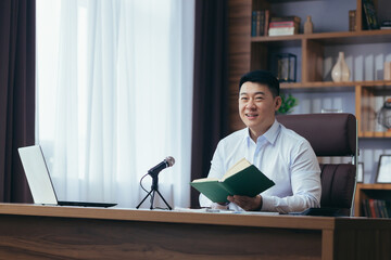 Asian preacher reads the bible online, records a podcast, sits at a table and looks at the camera, uses a microphone, holds his hand in his heart, online radio broadcast