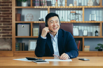 Asian businessman happy talking on video call in classic office, working at desk, using headset,...