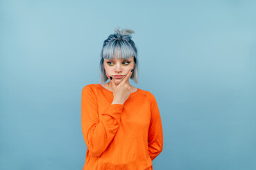 Pensive lady in orange sweater and blue hair isolated on blue background with serious face looking...
