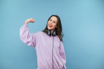 Attractive brunette girl in purple sweater and headphones shows biceps to camera and smiles.