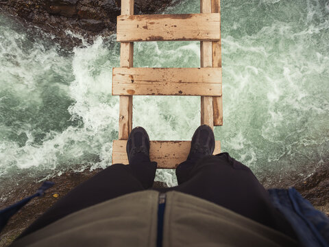 Men's feet stand on a narrow wooden bridge across a turbulent icy river. Optical distortion. View from above. Extreme river crossing